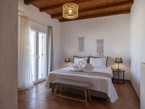 A bedroom with a bed, two bedside lamps, a bench, a hanging macrame wall hanging, and a wooden ceiling. - Myrsini Traditional Hillside Cottage (Paros)