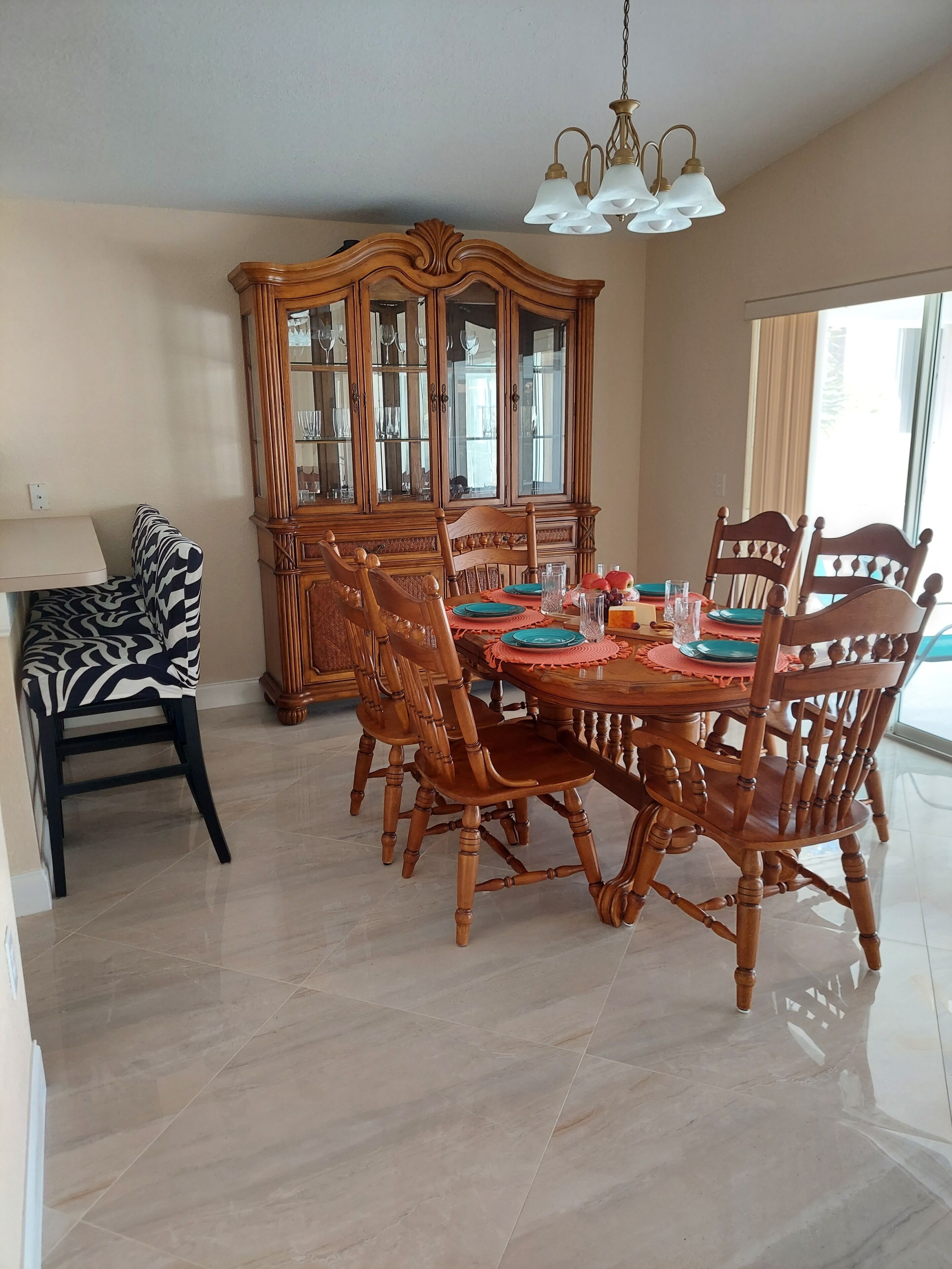 Uma sala de jantar com uma mesa de madeira preparada para quatro pessoas, rodeada por cadeiras a combinar. Um armário de madeira grande com portas de vidro está ao fundo.