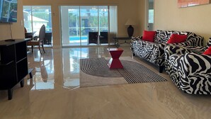 A spacious living room with a large sofa, a coffee table, and a TV. There is a ceiling fan and a window with a view of the outside. - HEATED POOL-LANAI-BBQ-5 Mi to BEACH-BICYCLES-TENNIS-GOLF-BLISSFUL RETREAT (Rotonda West)
