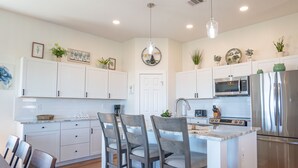 A modern kitchen with white cabinets, a central island with bar stools, stainless steel appliances, and decorative plants. - Beautiful Beachfront Home with Beach Access. (Sargent)