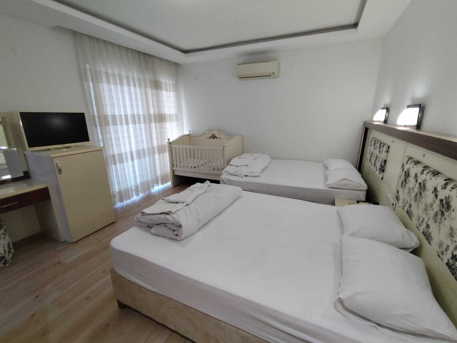A hotel room with two single beds, a desk with a television, a small white cabinet, and a window with sheer curtains.