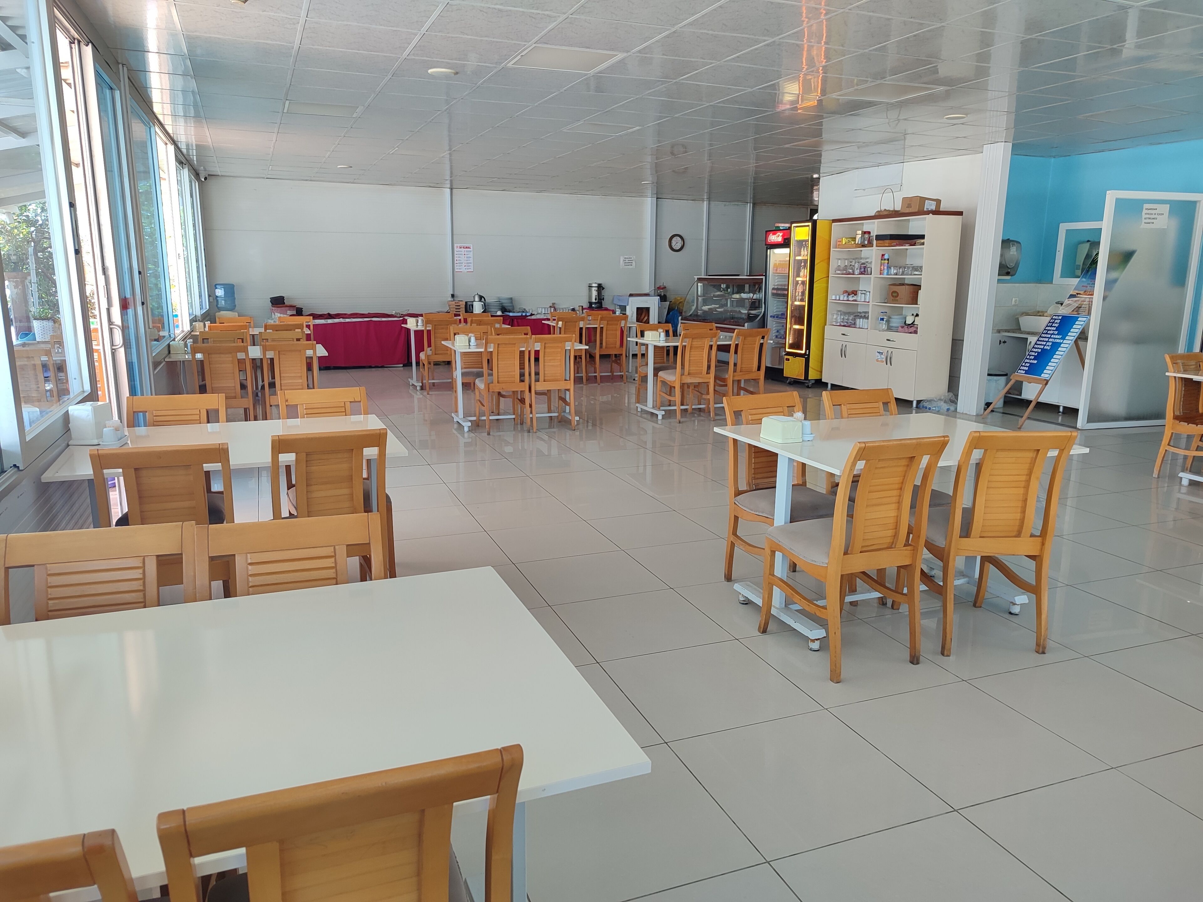 A spacious cafeteria with wooden tables and chairs, a vending machine, and a refrigerator.