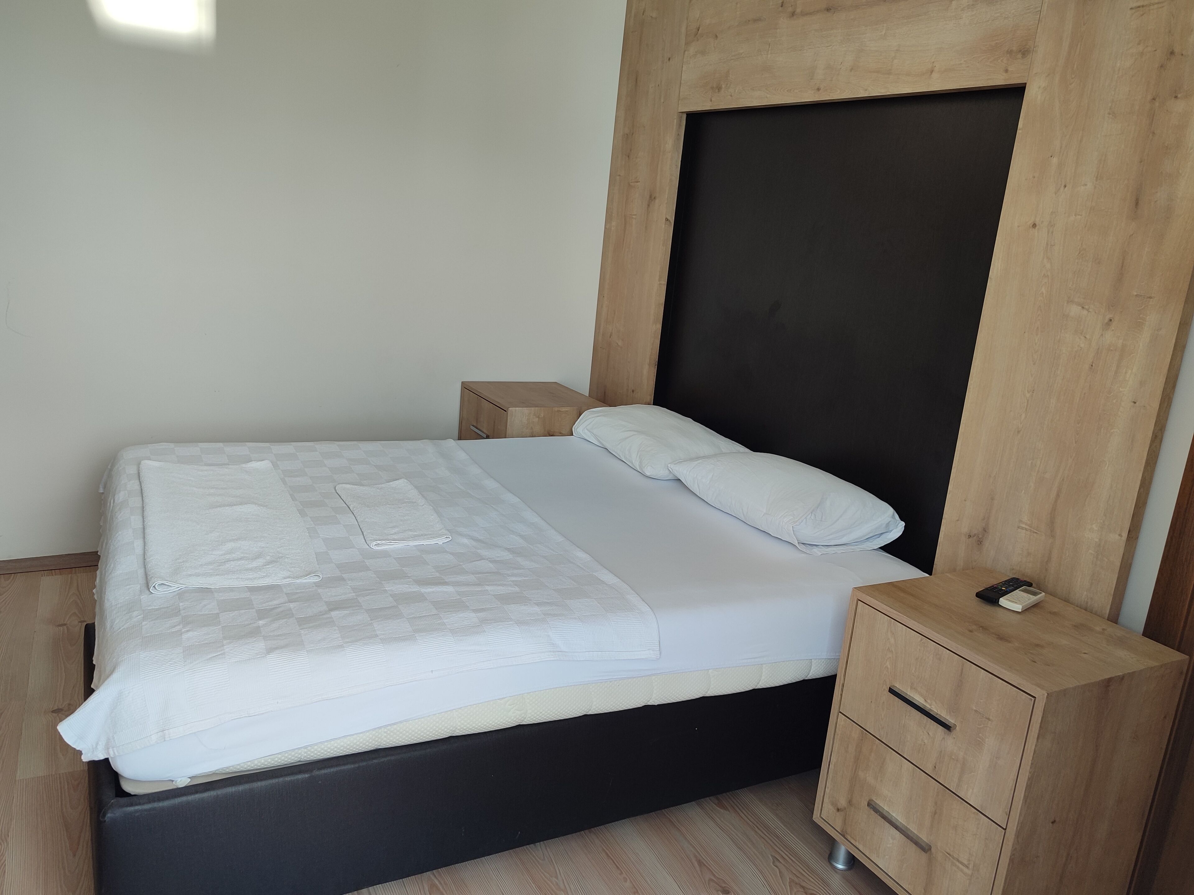 A neatly made bed with a checkered blanket and pillows, a wooden nightstand with drawers, and a dark headboard.