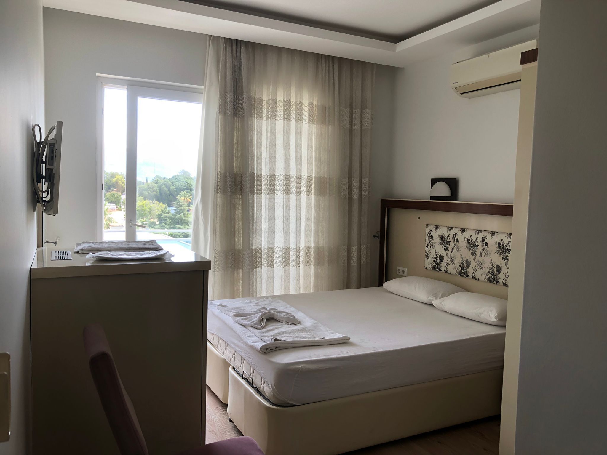 Habitación de hotel con cama, escritorio con silla, ventana con cortinas y aire acondicionado.