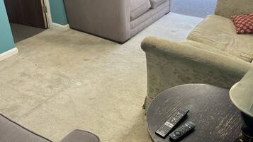 A living room with a beige sofa, a round table with two remote controls, and a view of an outdoor patio area.