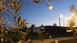 A campsite with a white RV parked on a gravel path, surrounded by trees and string lights. - Campeggio in Fattoria (Serre)