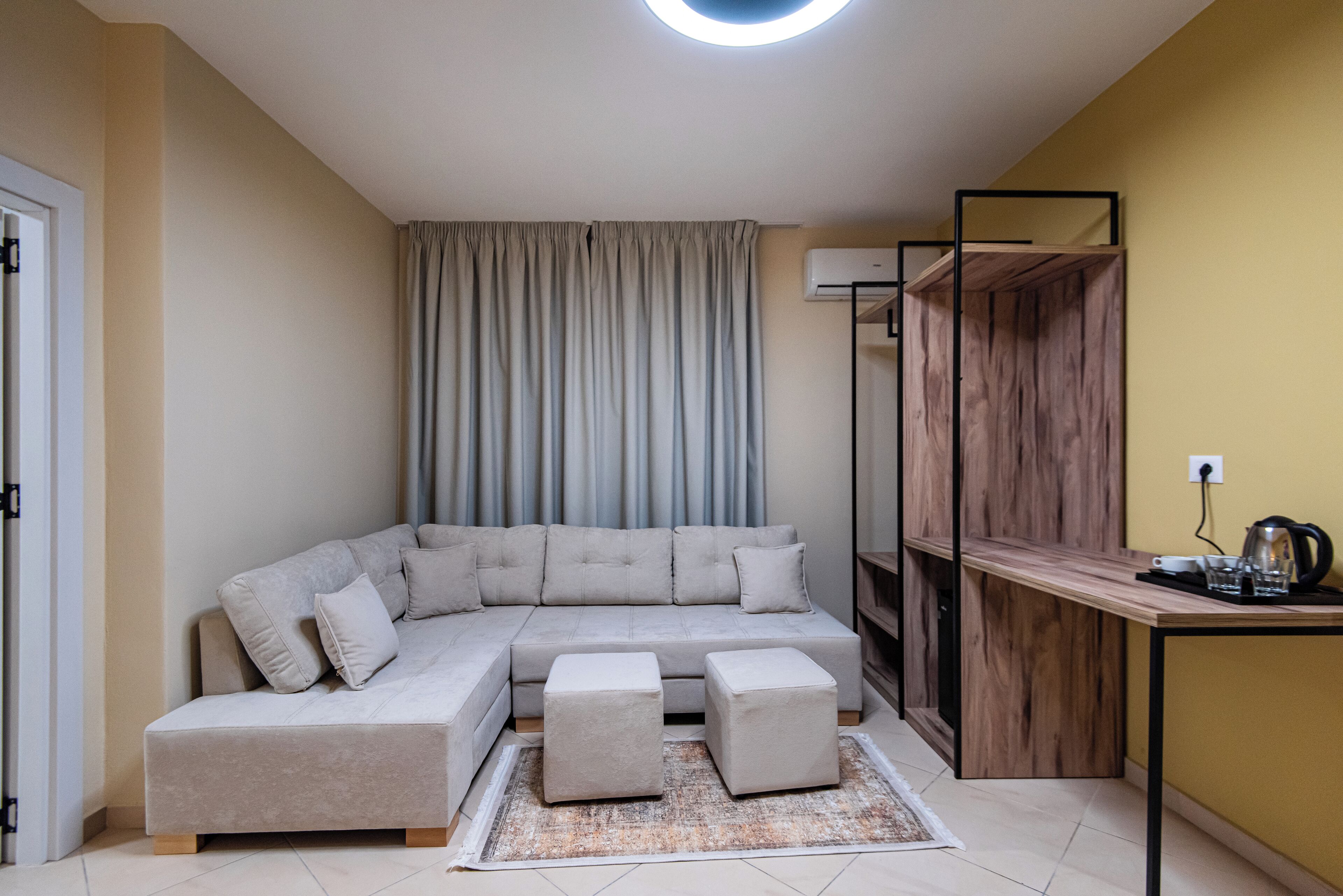 Un salon moderne avec un canapé d’angle, deux poufs, une table basse en bois et un miroir.