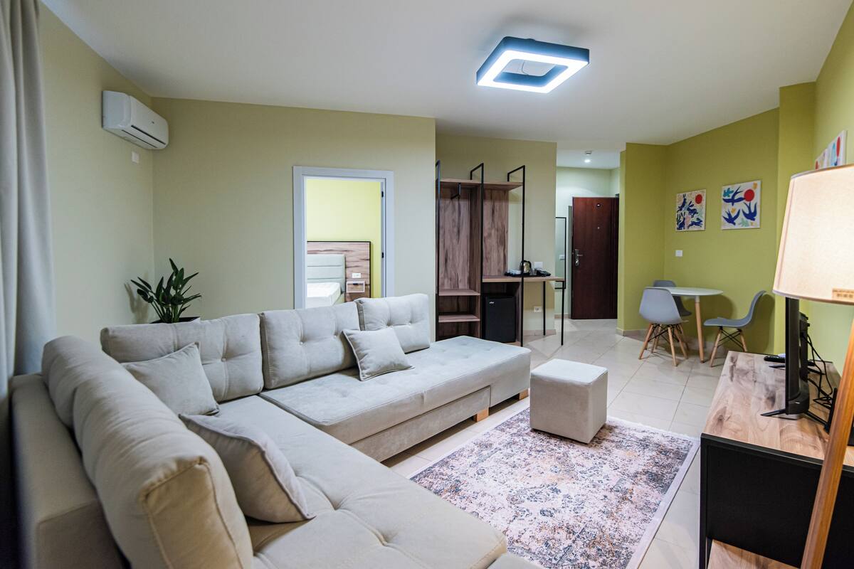 A living room with a sofa, ottoman, and a small table with a lamp. There is a plant, a ceiling light, and a door leading to another room.