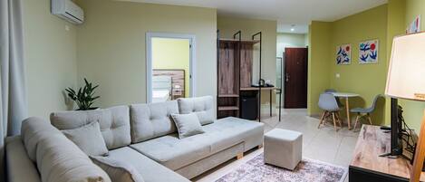 A living room with a sofa, ottoman, and a small table with a lamp. There is a plant, a ceiling light, and a door leading to another room.