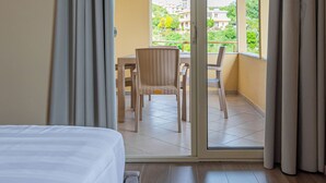 Habitación de hotel con cama, balcón con mobiliario de mimbre y vistas a zonas verdes y edificios.