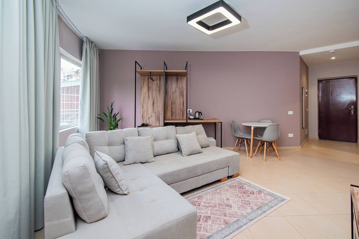A modern living room with a grey sectional sofa, a small dining table with chairs, a potted plant, and a patterned rug.