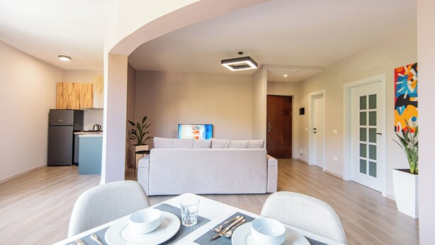 Una sala de estar moderna con sofá, mesa de comedor para dos personas y zona de cocina con refrigerador y planta.
