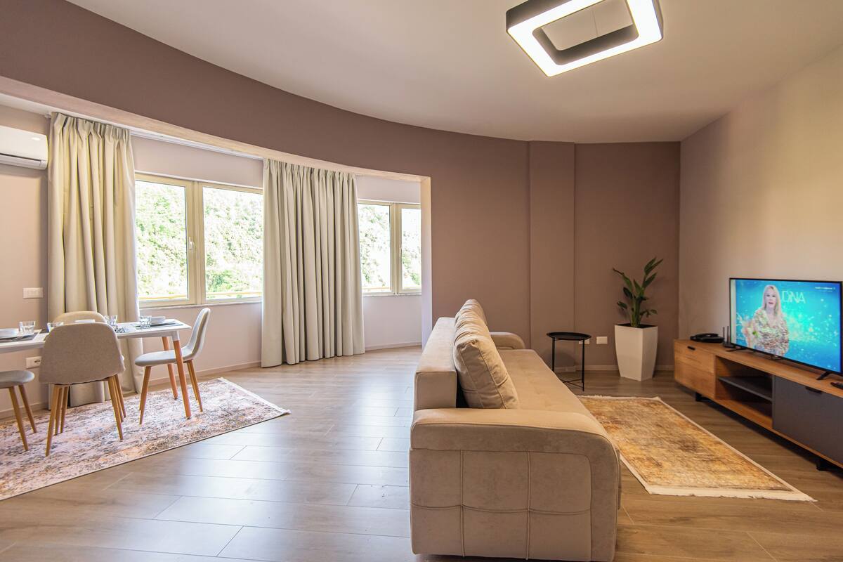 A modern living room with a sofa, TV, dining area, and large windows.