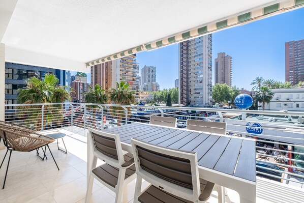 Un balcón con mesa y sillas, con vista a un paisaje urbano con edificios altos y palmeras.