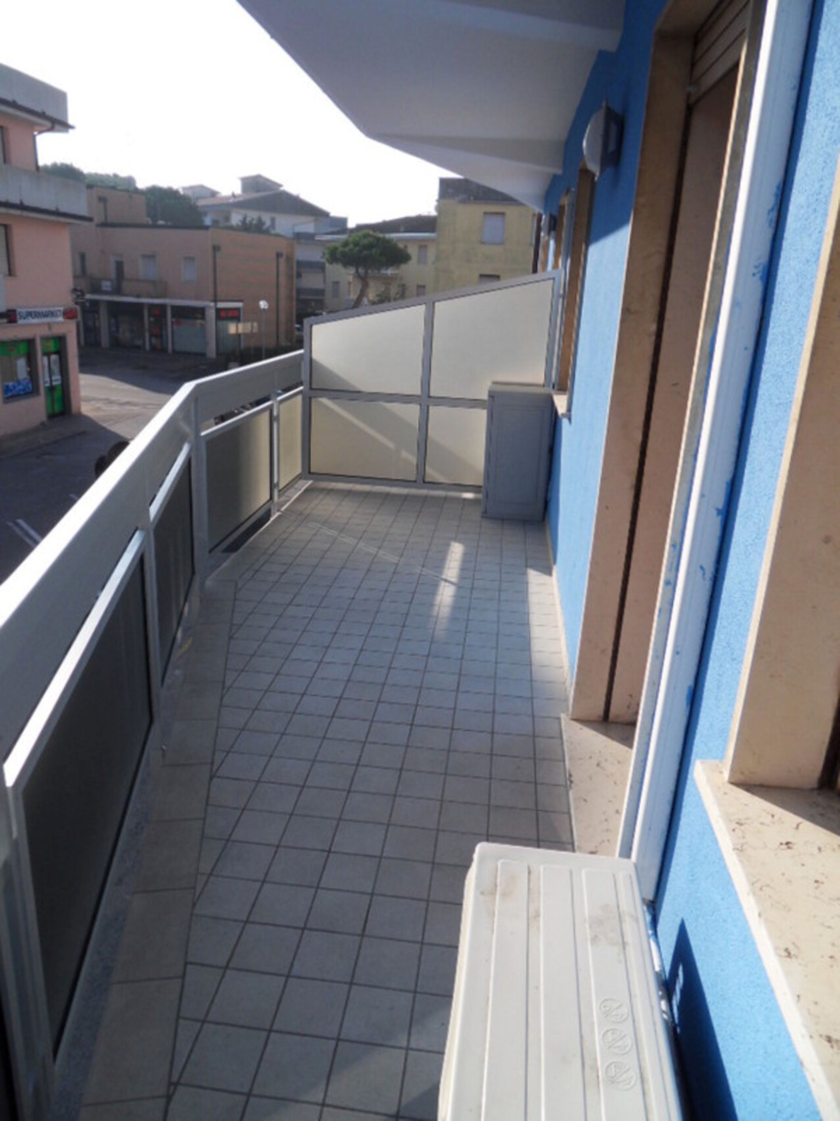 Uma varanda com chão de azulejos, um banco branco e vista para a rua e edifícios.