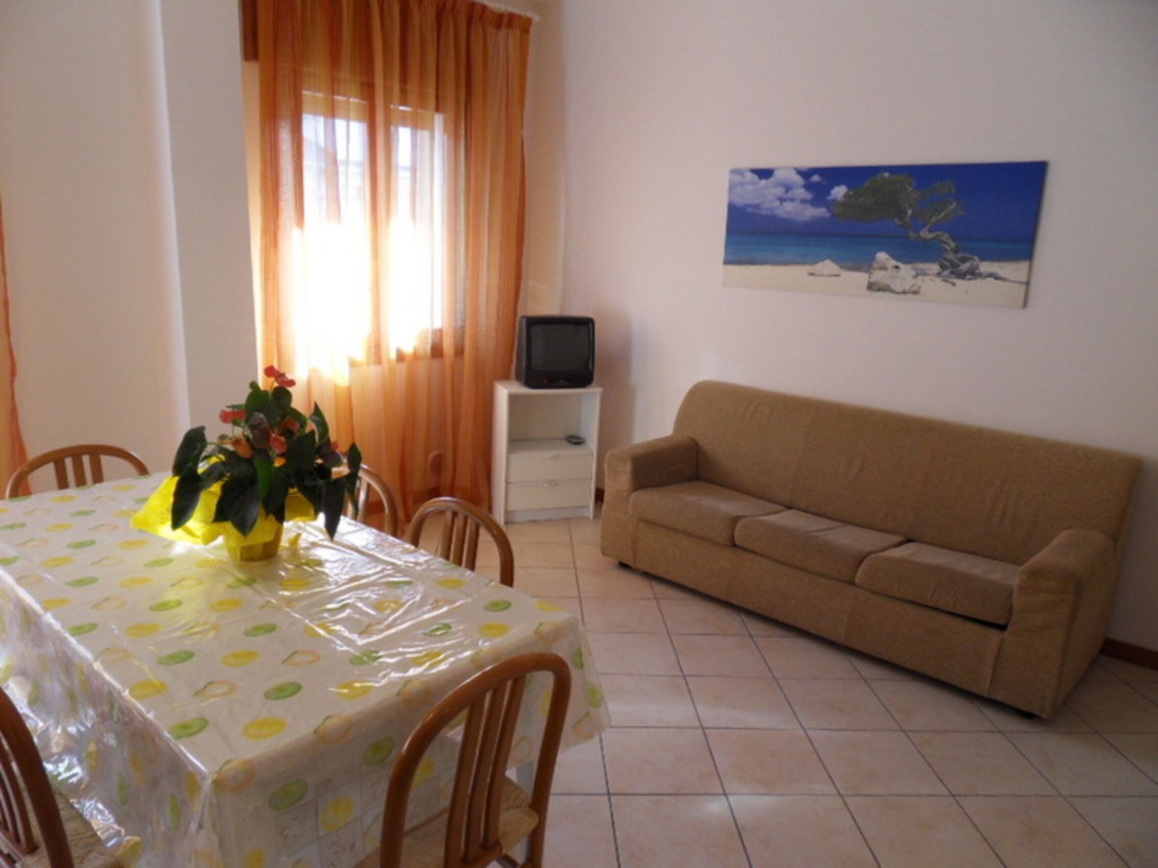 Uma sala de estar com um sofá bege, uma mesa de jantar com toalha de mesa de bolinhas e um quadro de uma cena de praia na parede.