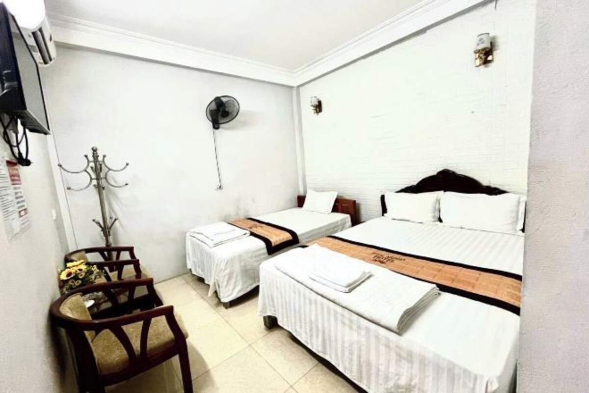 A hotel room with two beds, a desk, a chair, a fan, and a wall-mounted light fixture.