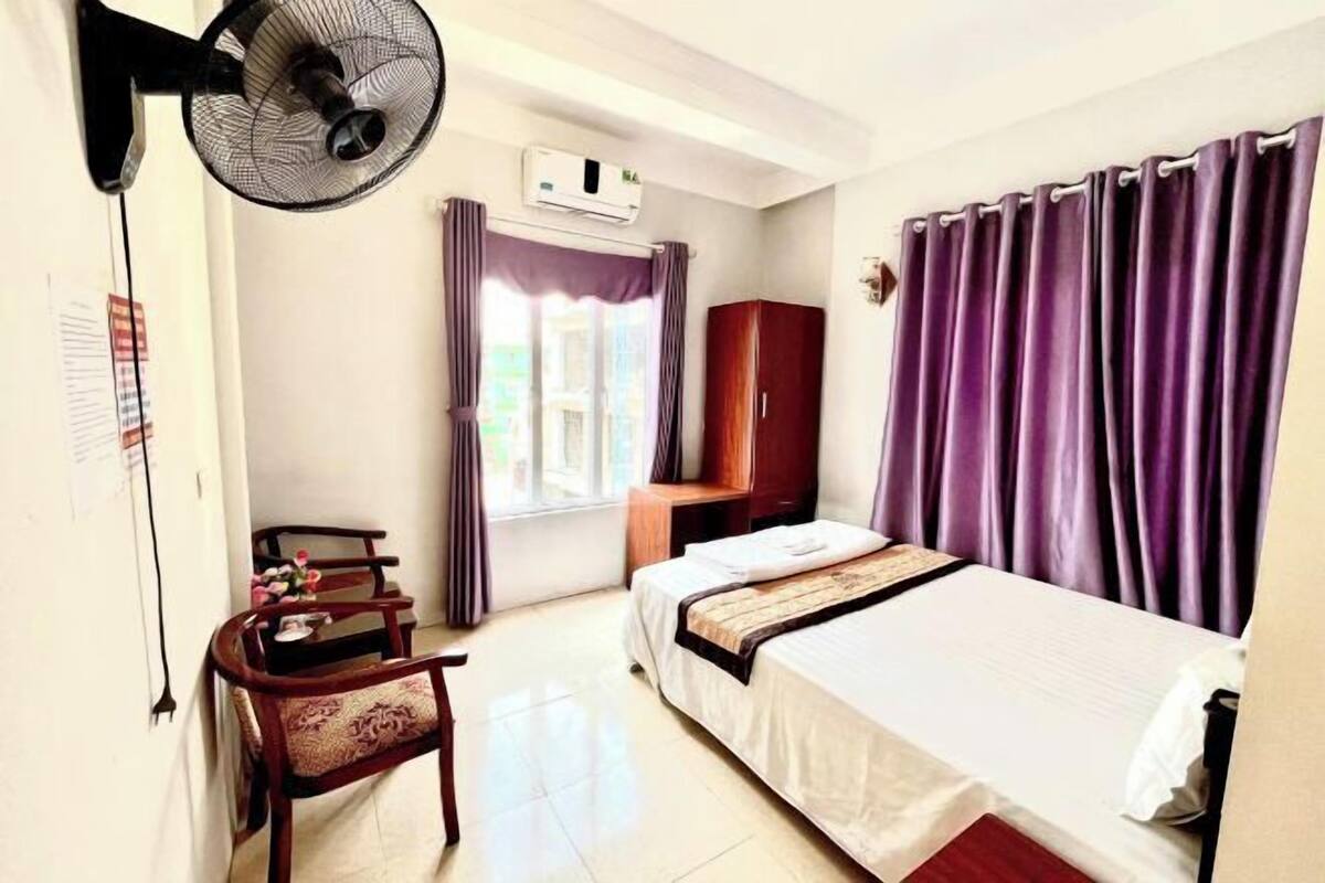 Habitación de hotel con cama, escritorio, silla, ventilador de pie, aire acondicionado y ventana con cortinas.