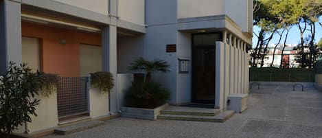 Un edificio moderno con entrada protegida por un portón, un pequeño jardín con plantas y un patio empedrado.