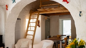 Ein gemütliches Zimmer mit einer Holzleiter, weißen Stühlen, einem Esstisch und Blick durch einen rundbogigen Durchgang.