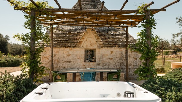 Ein Steinhaus mit einem kegelförmigen Dach, eine Holzpergola mit Weinreben und eine weiße Whirlwanne im Vordergrund.