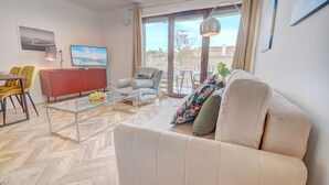 A modern living room with a sofa, armchairs, a glass coffee table, and a red cabinet. There is a TV mounted on the wall and a large window with curtains. - HelApart - Belnihel (Hel)
