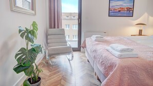 A hotel room with a bed, a chair, a potted plant, a painting, and a view of a building through the window. - HelApart - Belnihel (Hel)