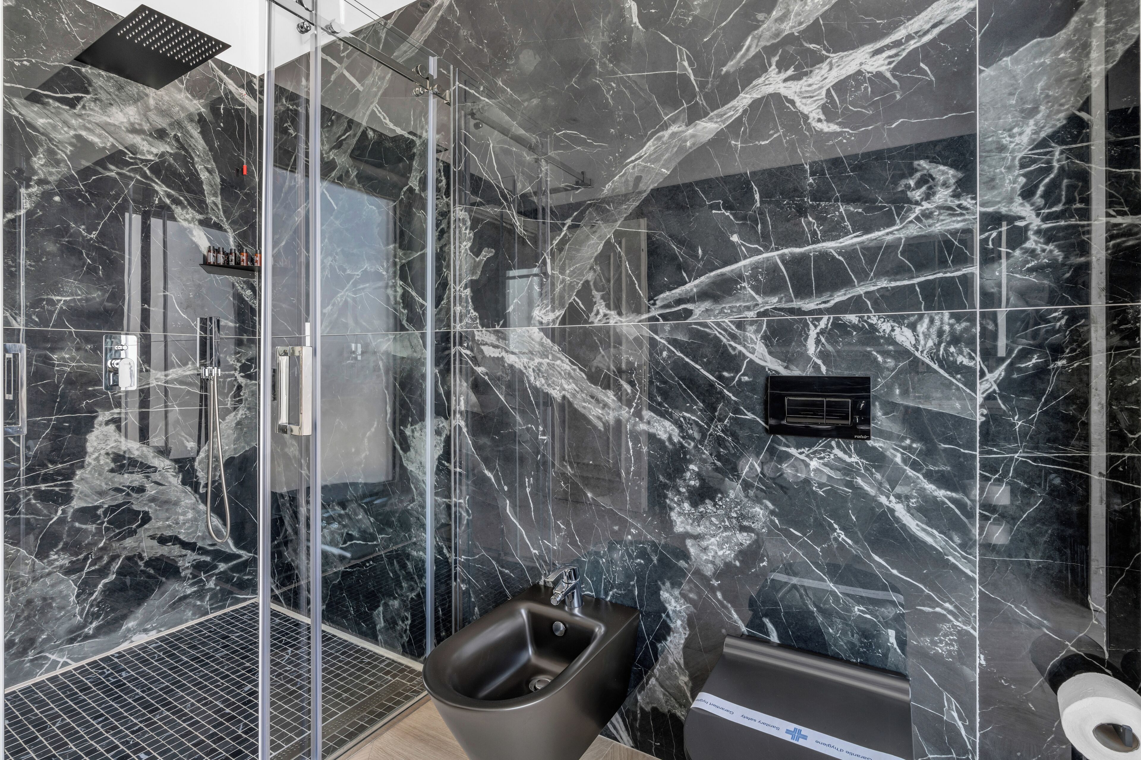 A modern bathroom with a glass shower enclosure, a bidet, and a marble-tiled wall.