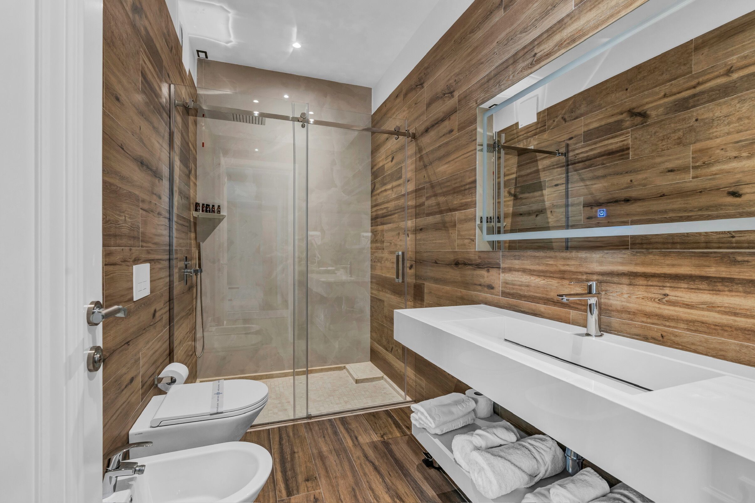 Un baño moderno con ducha envuelta en cristal, lavamanos blanco y paneles de madera.