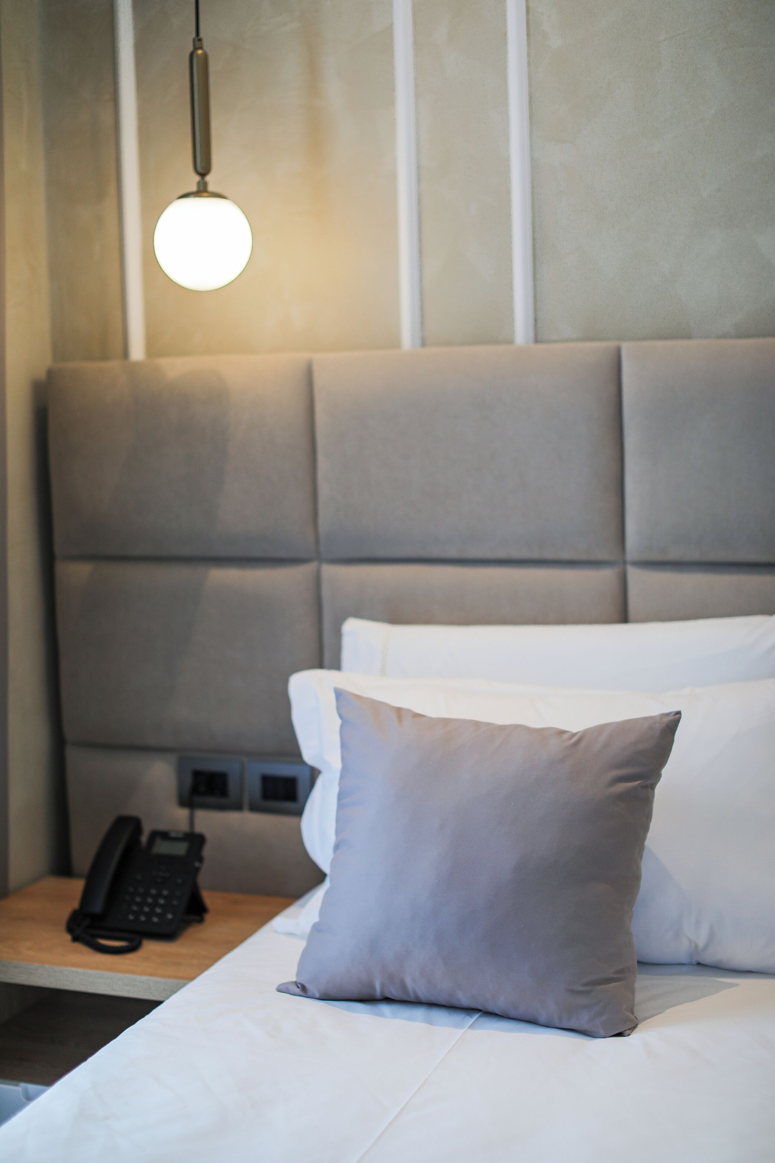 Una cama bien hecha con un almohadón gris, un teléfono en la mesita de noche y una lámpara colgante.