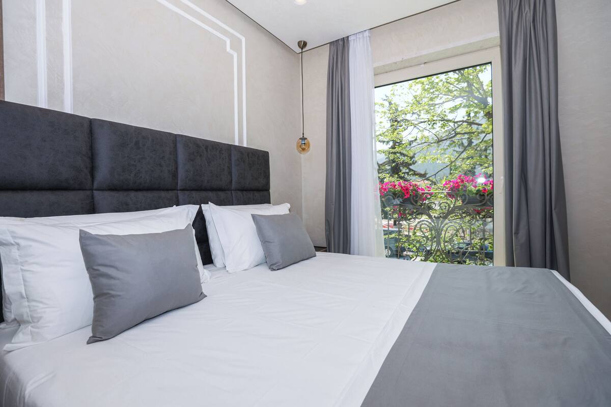 Una habitación de hotel moderna con una cama grande, ropa de cama gris y blanca, y vistas a un balcón con flores.