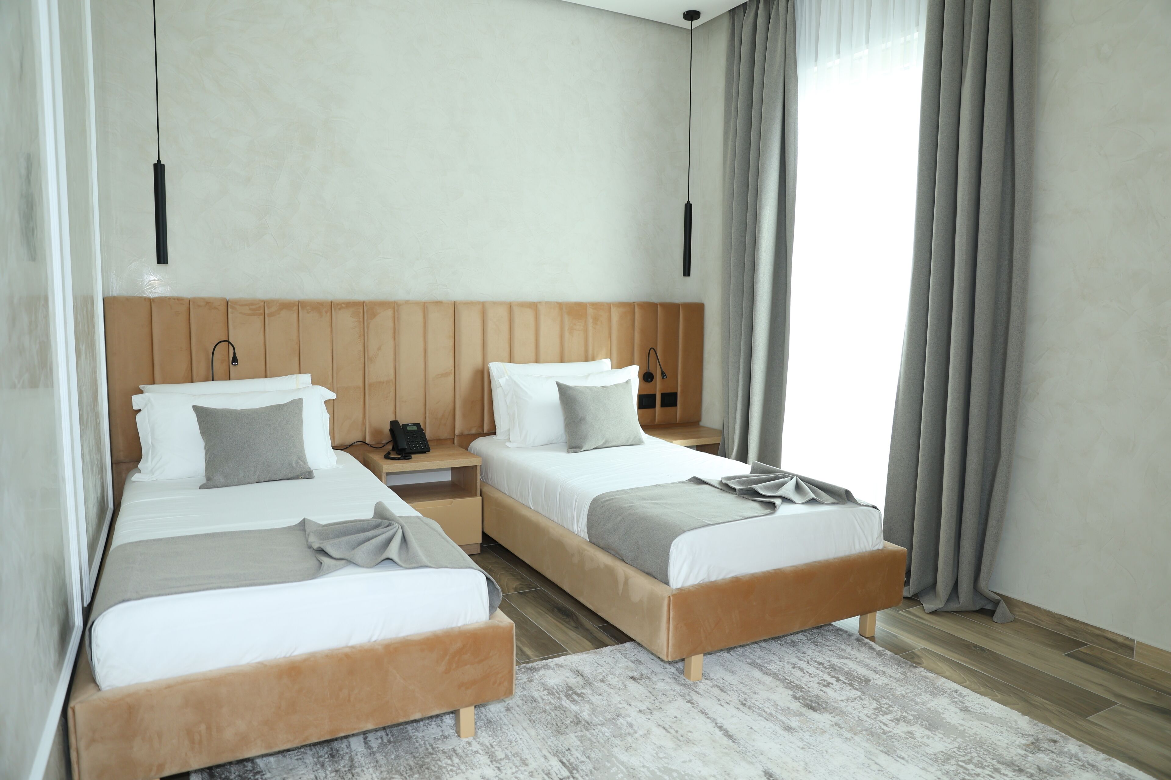 A modern hotel room with two beds, wooden headboards, a white and grey color scheme, and a large window with curtains.