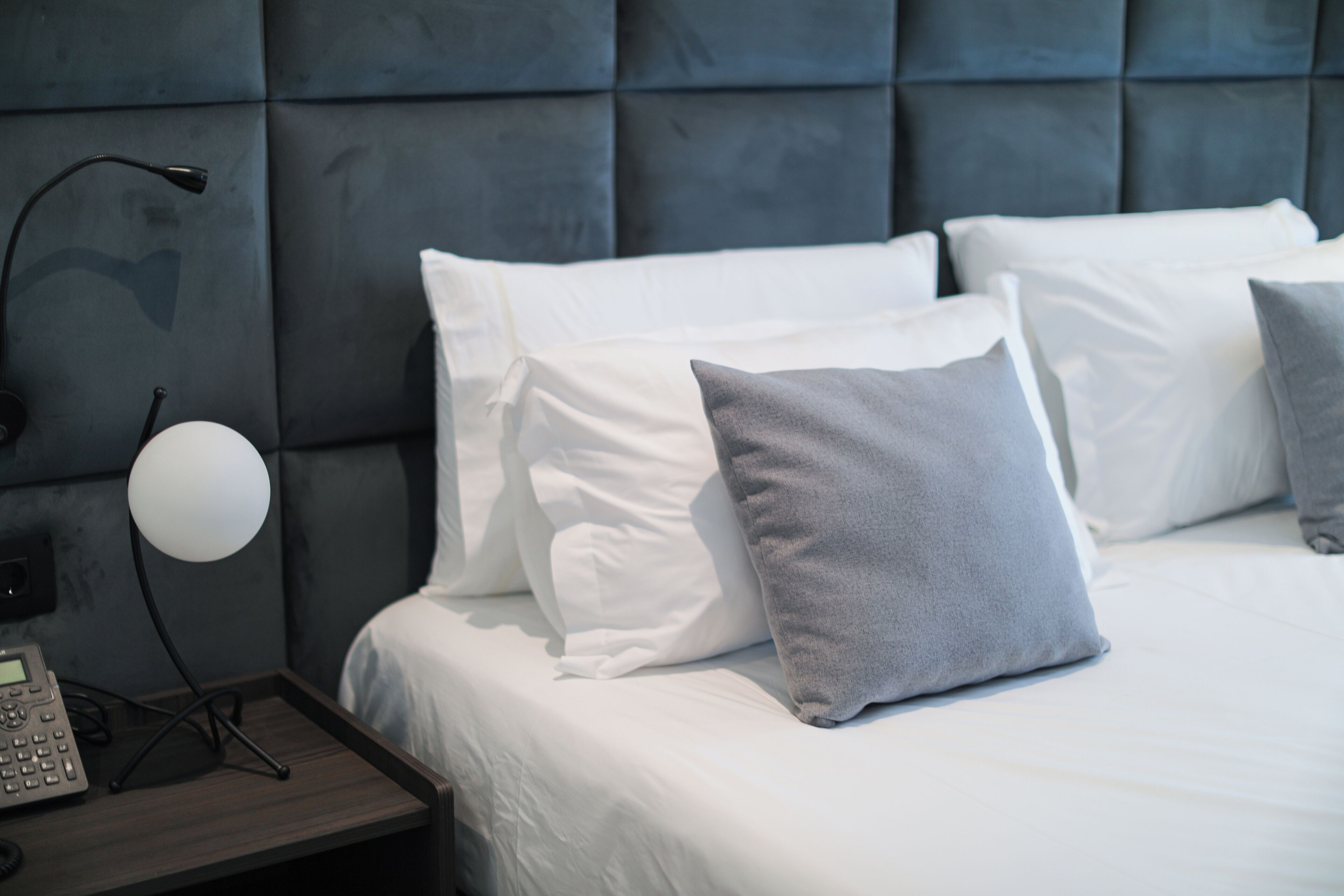 Een keurig opgemaakt bed met witte en grijze kussens, een bedlamp en een telefoon op een houten nachtkastje.