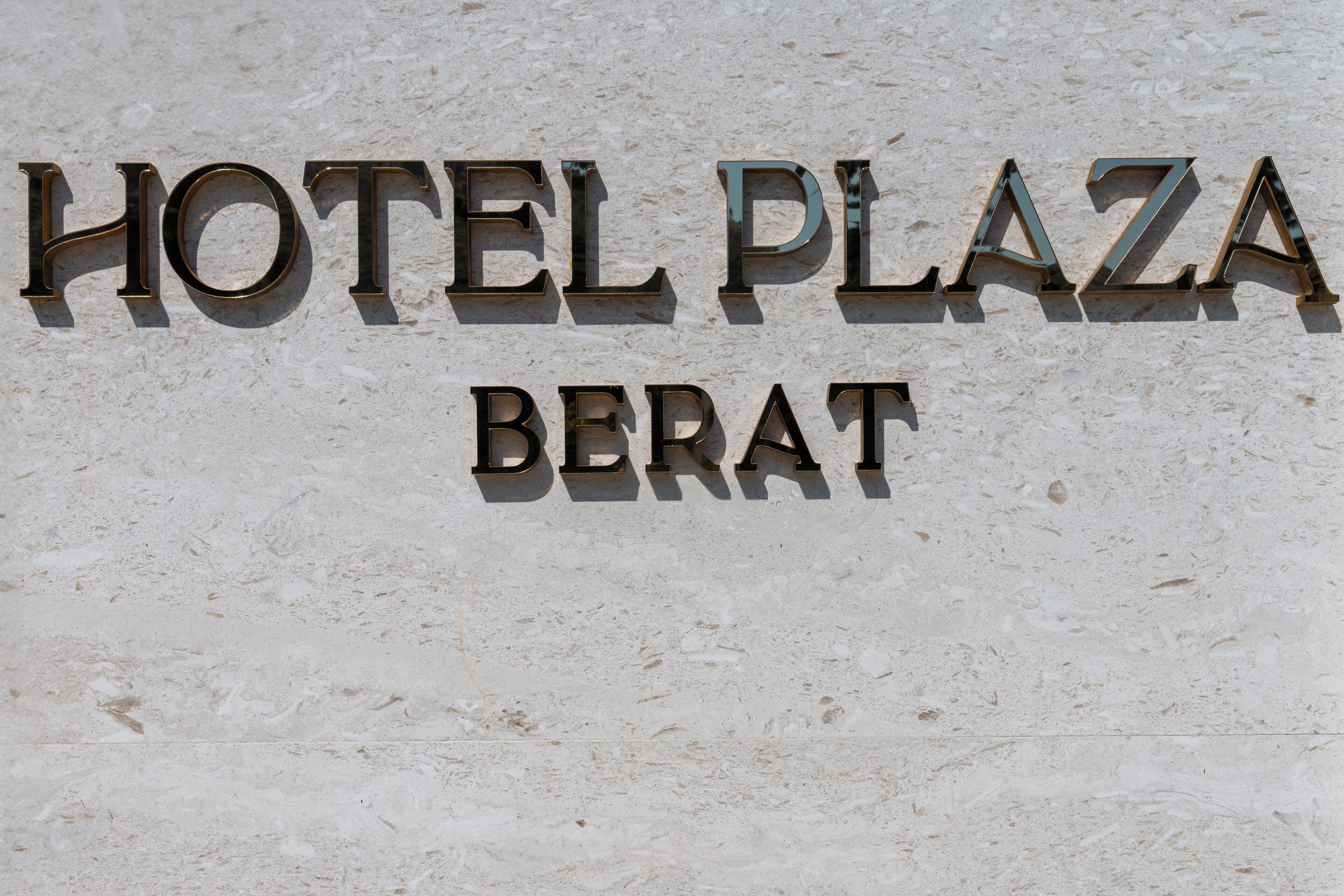 Ein Schild mit der Aufschrift 'HOTEL PLAZA BERAT' in Metallbuchstaben an einer strukturierten Wand.