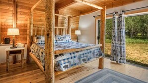 Ein Schlafzimmer einer Holzhütte mit einem Himmelbett, einem kleinen Tisch mit Lampe und Blick ins Freie durch ein großes Fenster.