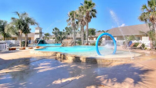 A pool area with a water feature, palm trees, and a building in the background. - Ground level condo near beach with a deck, washer/dryer AC & Pool (Corpus Christi)