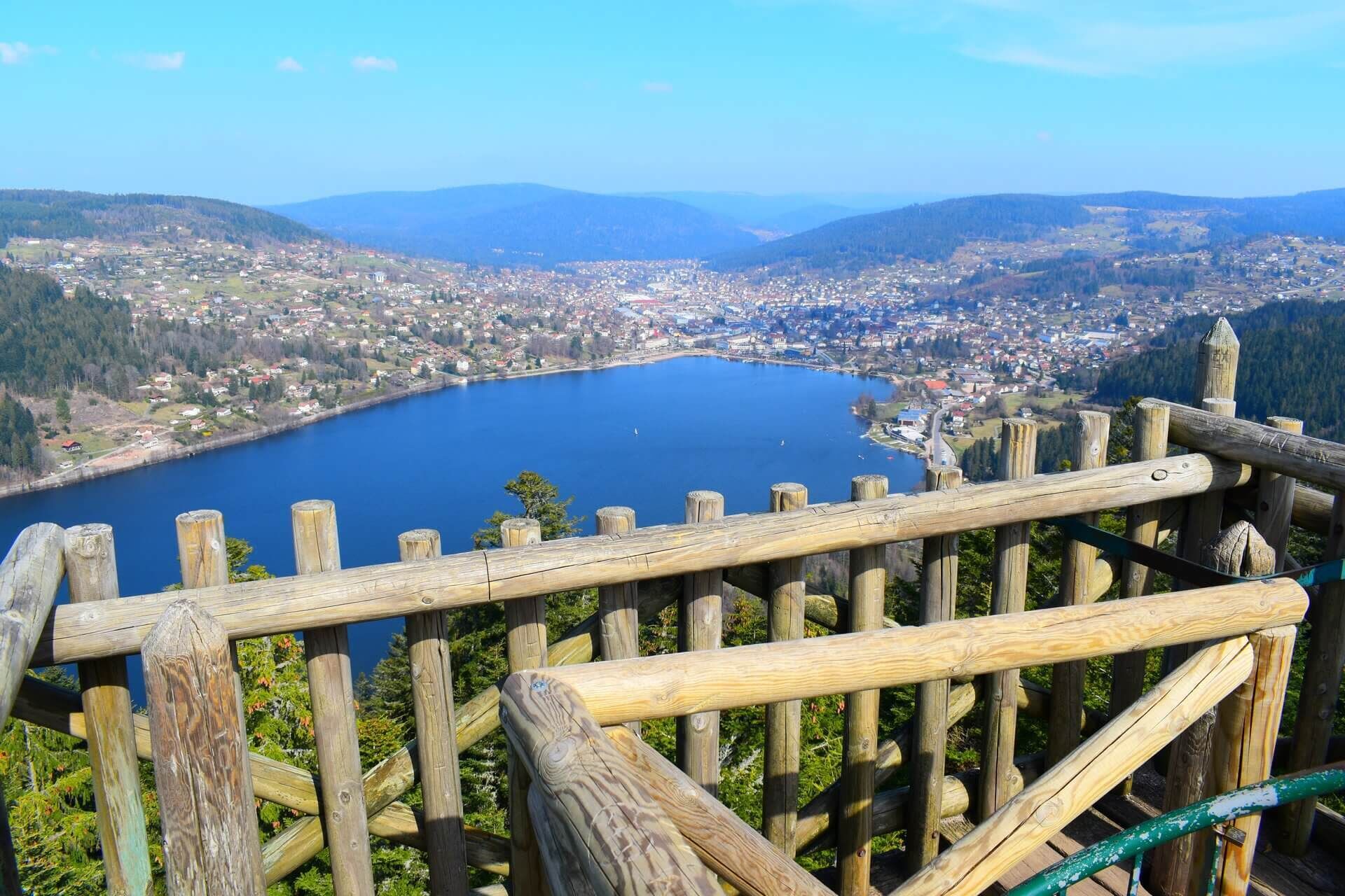Une vue panoramique d’un lac entouré d’une ville et de montagnes, prise depuis une plateforme d’observation en bois.