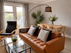 Un salon moderne avec un canapé en cuir, une table basse en verre, une table à manger avec des chaises, une lampe sur pied et une grande fenêtre avec des rideaux.