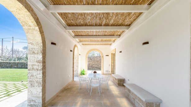 Ein überdachter Patio-Bereich mit Steinboden, weißen Wänden und einem zentralen Tisch mit Stühlen.