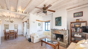 A spacious living room with a wooden ceiling, a stone fireplace, a white sofa, a dining table with chairs, and a bookshelf.