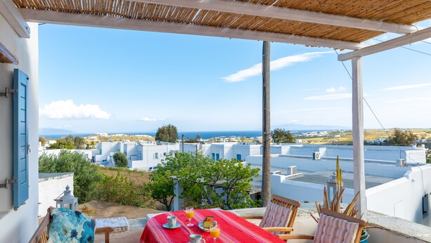 A terrace with a table set for breakfast, overlooking a coastal village with white buildings and a clear sky.