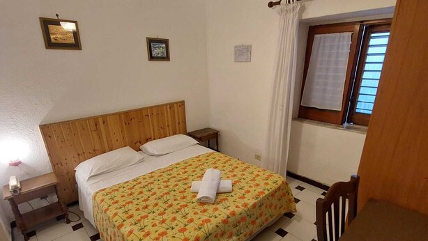 Habitació d'hotel amb llit de fusta, taula de nit petita, taulell, escriptori amb cadira, finestra amb cortines i lluminària penjada a la paret.