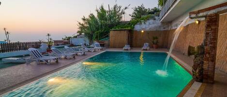 Una piscina con sillas de descanso, un muro de ladrillo y vistas a la puesta de sol.