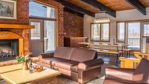 A cozy living room with a fireplace, leather sofa, wooden tables, and large windows. - Spacious 8 bdrm Blue Mountain Cottage, Perfect for Large Groups, Private Beach, BBQ, Hot Tub, Sauna (The Blue Mountains)