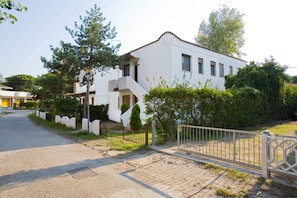 Edifício branco com varanda, portão e vegetação ao redor.
