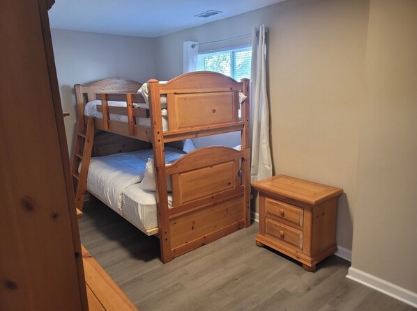 Una cama litera con cabecera de madera, una mesita de noche de madera y una ventana con cortinas.