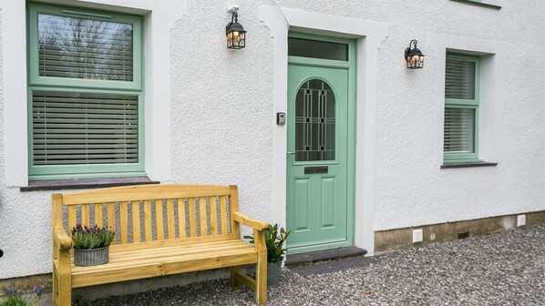 Un edificio blanco con una puerta verde, un banco de madera y dos ventanas con contraventanas verdes.