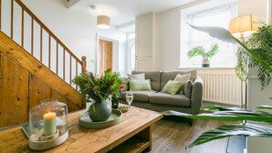 A cozy living room with a wooden staircase, a grey sofa, and a wooden coffee table with a candle, glass jar, and a vase of flowers. - Ty Capel -  a dog that sleeps 8 guests  in 4 bedrooms (Pentraeth)