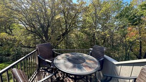 Une terrasse extérieure avec une table ronde en verre et des chaises en osier, entourée d’arbres.
