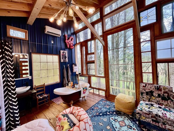 A rustic cabin interior with a large window, a bathtub, and a patterned rug. - Treehouse + Trolley Parton - 2 Unique Stays - Why Choose? (Doyle)